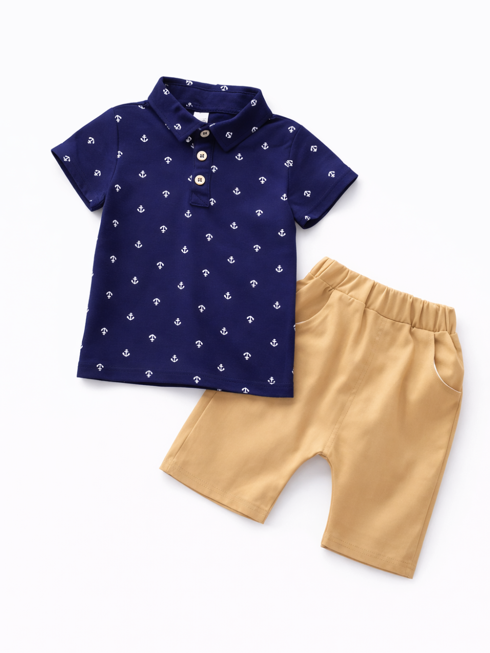 白背景のスタジオ撮影。ネイビーの半袖ポロシャツとベージュのショートパンツのキッズ2点セットが配置されている。ポロシャツは小さな白いいかり柄プリントと襟付きデザインで、木目調ボタン3つ付き。ショートパンツはウエストゴム仕様でサイドポケットがある。
