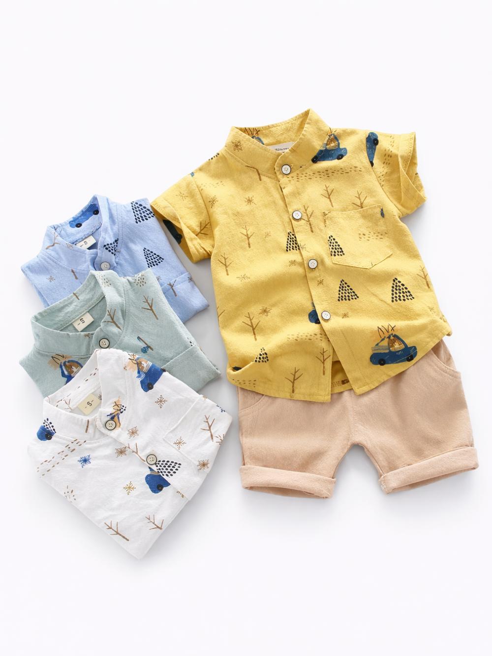 白背景のスタジオ撮影。子ども用の半袖シャツとショートパンツのセットが1組（黄色シャツ＋ベージュショーツ）と、折りたたまれた替えシャツ3枚（ライトブルー、ミント、ホワイト）が並んで配置されている。シャツは木や三角形の柄、クリスマスツリーを載せた車のイラストなどのプリント入りで、前ボタンと胸ポケット付き。ショートパンツは裾を折り返したデザイン。

