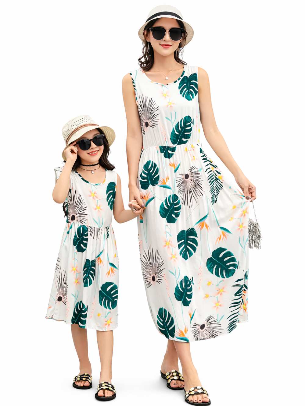 白地のスタジオ背景で、トロピカル柄（グリーンの葉と花）の白いワンピースを着た母親と女の子が手をつないで立っている。母親はノースリーブのロング丈、女の子はノースリーブのひざ丈で、どちらも帽子と黒いサングラス、サンダルを着用している。
