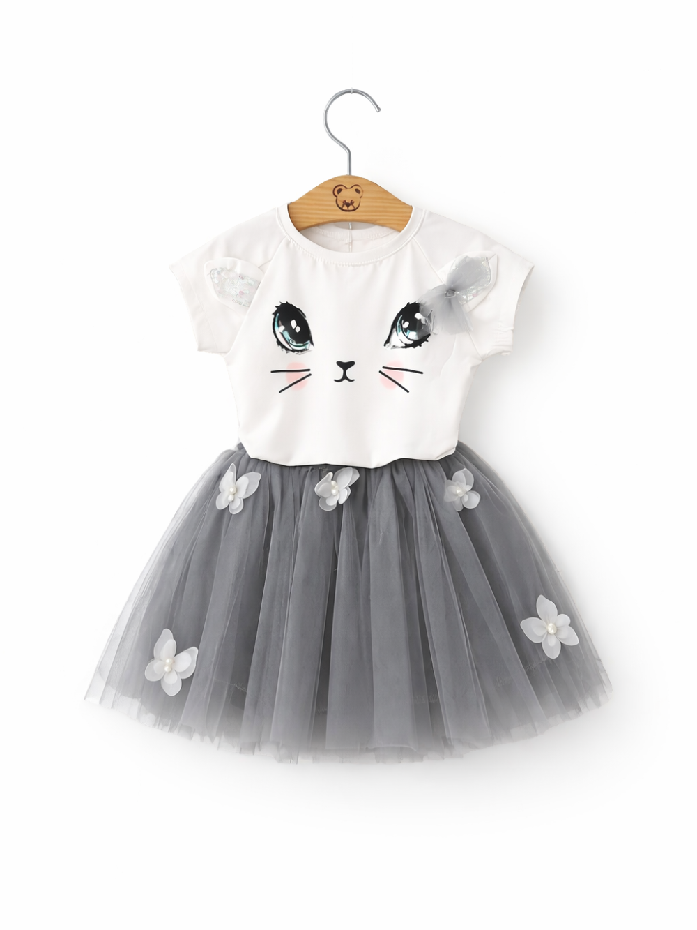 女の子用ワンピース。白い半袖トップスに猫の顔プリント（大きな目・ひげ・ほほ色）と肩のリボン付き。グレーのチュールスカートは立体の花飾り付き。ハンガーに掛けた状態で白背景。

