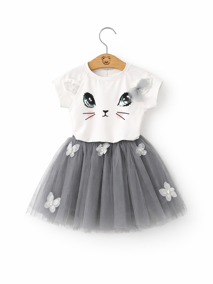 女の子用ワンピース。白い半袖トップスに猫の顔プリント（大きな目・ひげ・ほほ色）と肩のリボン付き。グレーのチュールスカートは立体の花飾り付き。ハンガーに掛けた状態で白背景。
