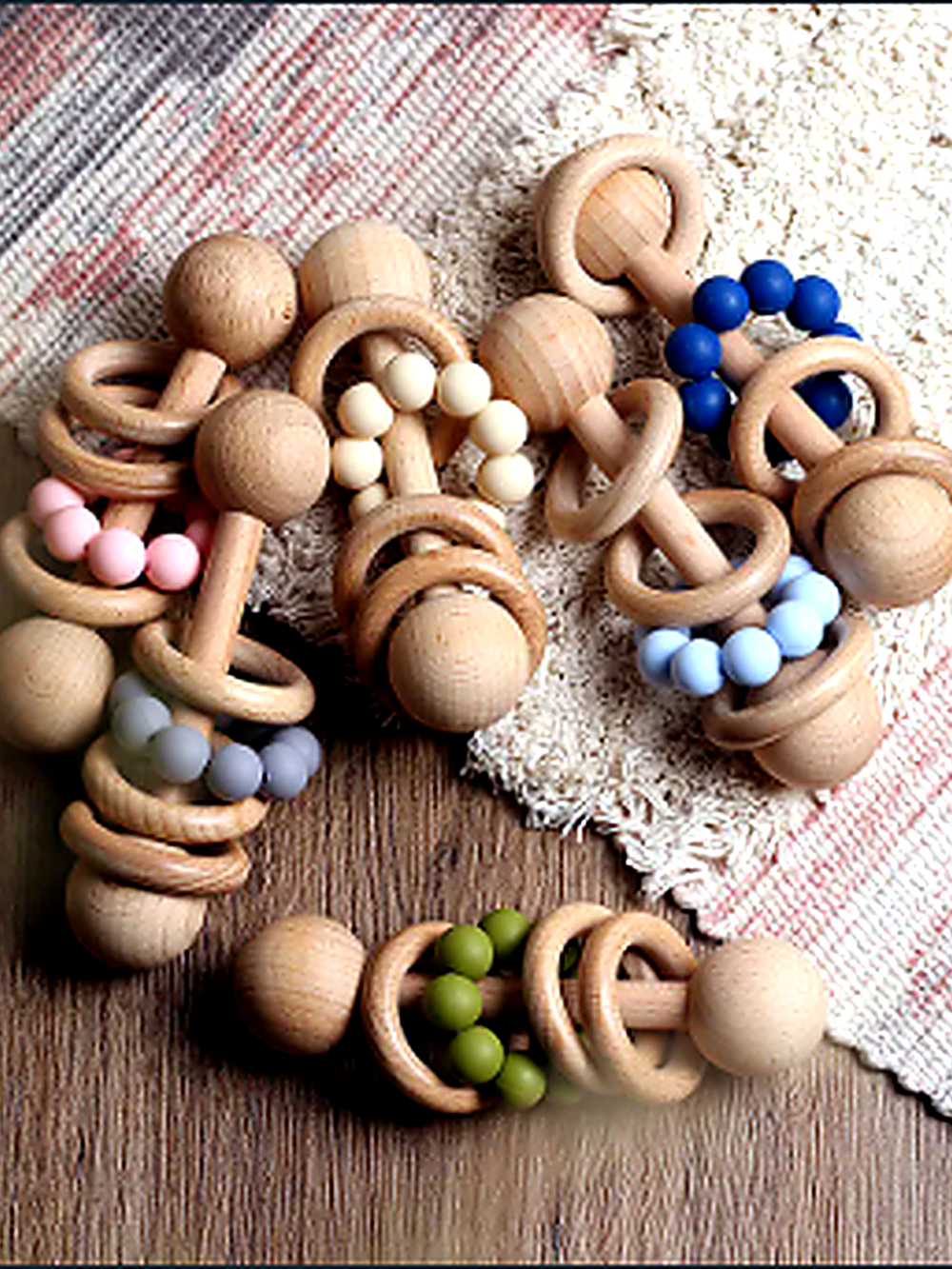 木目調の床とラグの上に、天然木のベビー用ガラガラ（リング付きラトル）を複数並べた商品画像。木製リングとビーズ（ピンク／ブルー／グリーンなど）の組み合わせで、ナチュラルな質感とカラーバリエーションが分かる。
