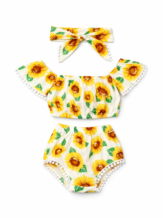 ひまわり柄のキッズ2点セット（トップス＋ショーツ）とリボンヘアバンド。クリーム地に黄色のひまわりとグリーンの葉のプリントで、袖口と裾に白いポンポンレース付き。白背景で単品。
