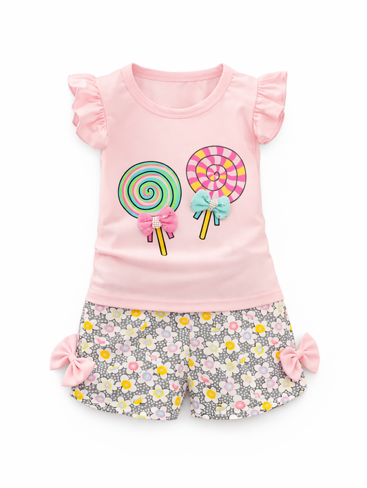 ピンクのフリル袖Tシャツと花柄ショートパンツの女の子用2点セット。トップスはカラフルなキャンディ（ロリポップ）プリントとリボン飾り付きで、パンツは両サイドにピンクのリボンが付いている。白背景で単品。
