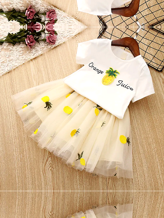 白いTシャツに「Orange Juice」の文字とパイナップルのアップリケが付いた子ども用ツーピースドレス。クリーム色のチュールスカートに黄色いパイナップル刺繍が散りばめられ、木目調の床の上にハンガーで平置きされている。左上にレース布とピンクのバラ、右上にワイヤーバスケットが写っている。
