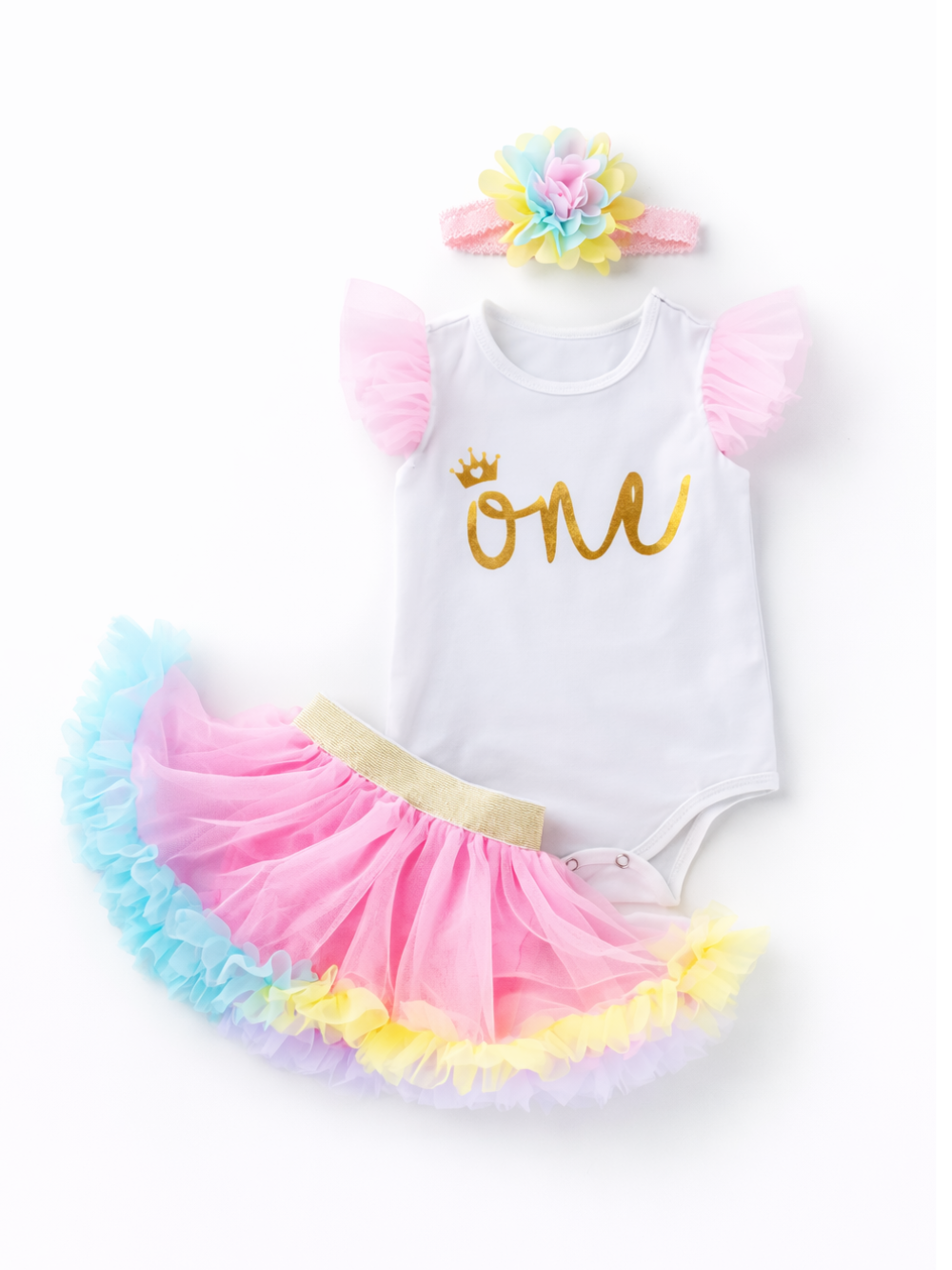乳児女の子用の1歳バースデー衣装を白背景でフラットレイ撮影した写真。ゴールドの「one」プリントと王冠モチーフの白いロンパース、ピンクのチュール袖、ゴールドウエストのピンク×ブルー×イエローのチュチュスカート、カラフルな花付きヘアバンドが並んでいる。
