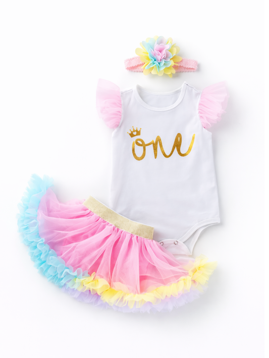 乳児女の子用の1歳バースデー衣装を白背景でフラットレイ撮影した写真。ゴールドの「one」プリントと王冠モチーフの白いロンパース、ピンクのチュール袖、ゴールドウエストのピンク×ブルー×イエローのチュチュスカート、カラフルな花付きヘアバンドが並んでいる。
