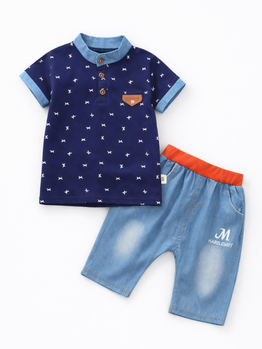 白い背景に平置きされた子ども用上下セット。小さな白柄のネイビーのポロシャツ（デニム風の襟・袖口、ボタン付き）と、オレンジのウエストバンドが付いたライトブルーのデニムショーツ。