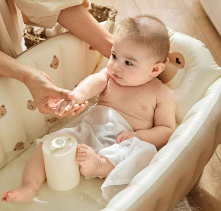ベージュの空気式ベビーバスの中で赤ちゃんがお湯につかり、大人がカップでやさしくお湯をかけている沐浴シーン（室内用インフレータブルベビーバス）
